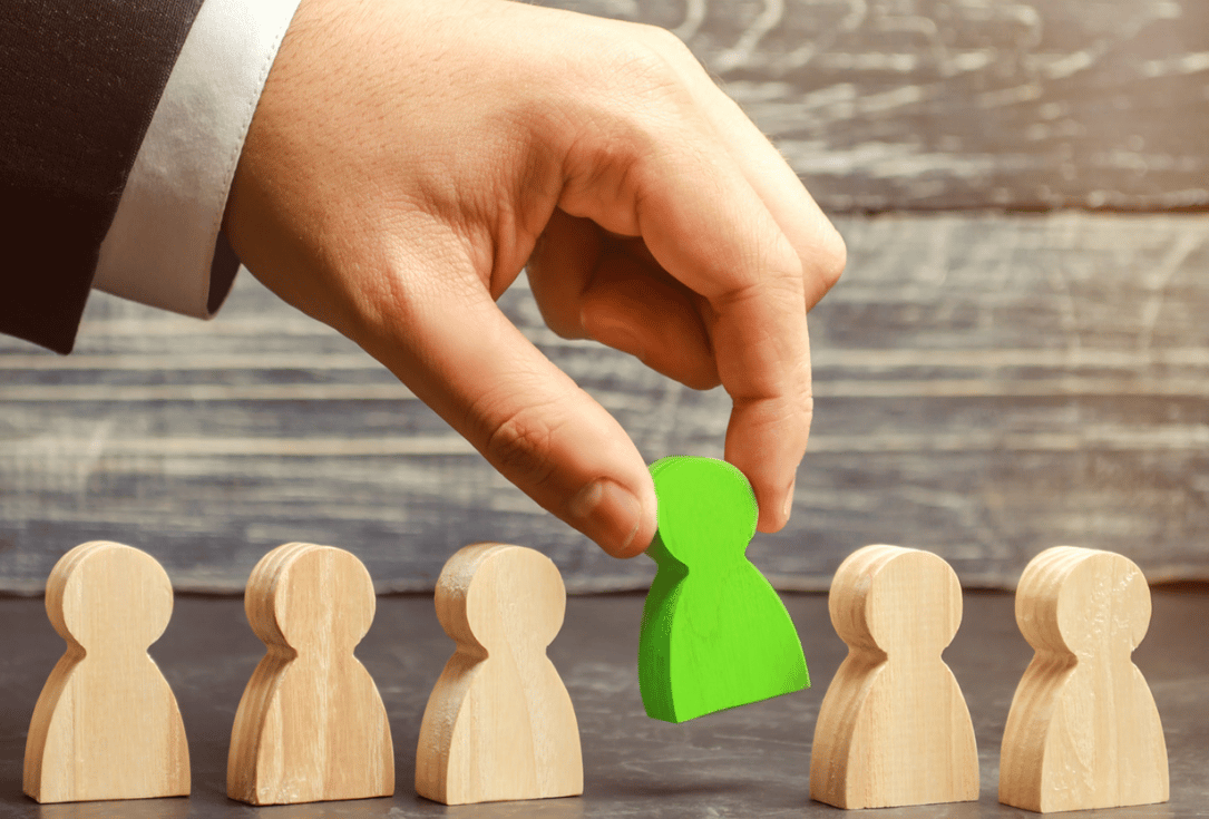 Hand places a green figurine among wooden figures to highlight a chosen candidate in a lineup of people at a recruitment or selection moment.