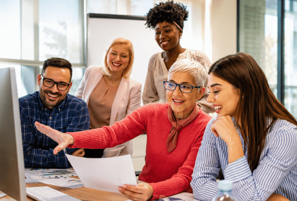 Five coworkers in a bright office review a document on a computer screen as one woman points to it—collaborative teamwork in action.