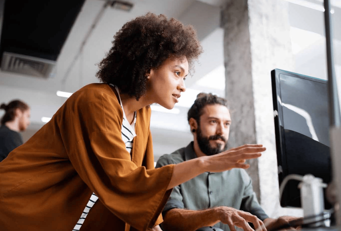 Two coworkers collaborate at a computer: a woman in a brown jacket points at the monitor while a man with a beard watches from the side in a bright office.