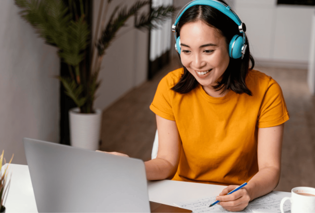Woman with teal headphones working at a desk, typing on a laptop and writing in a notebook.