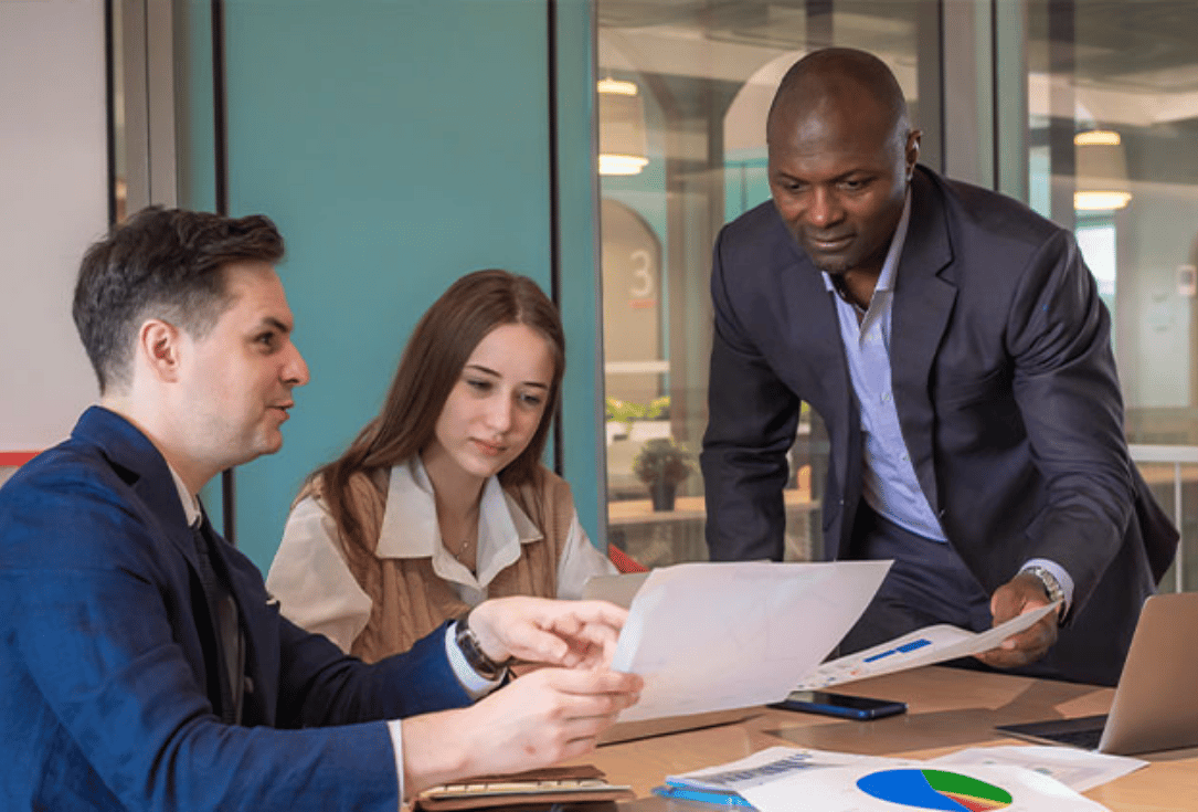 Three professionals in a meeting around a table reviewing documents; one points at a chart on paper while another holds a sheet and a laptop nearby.