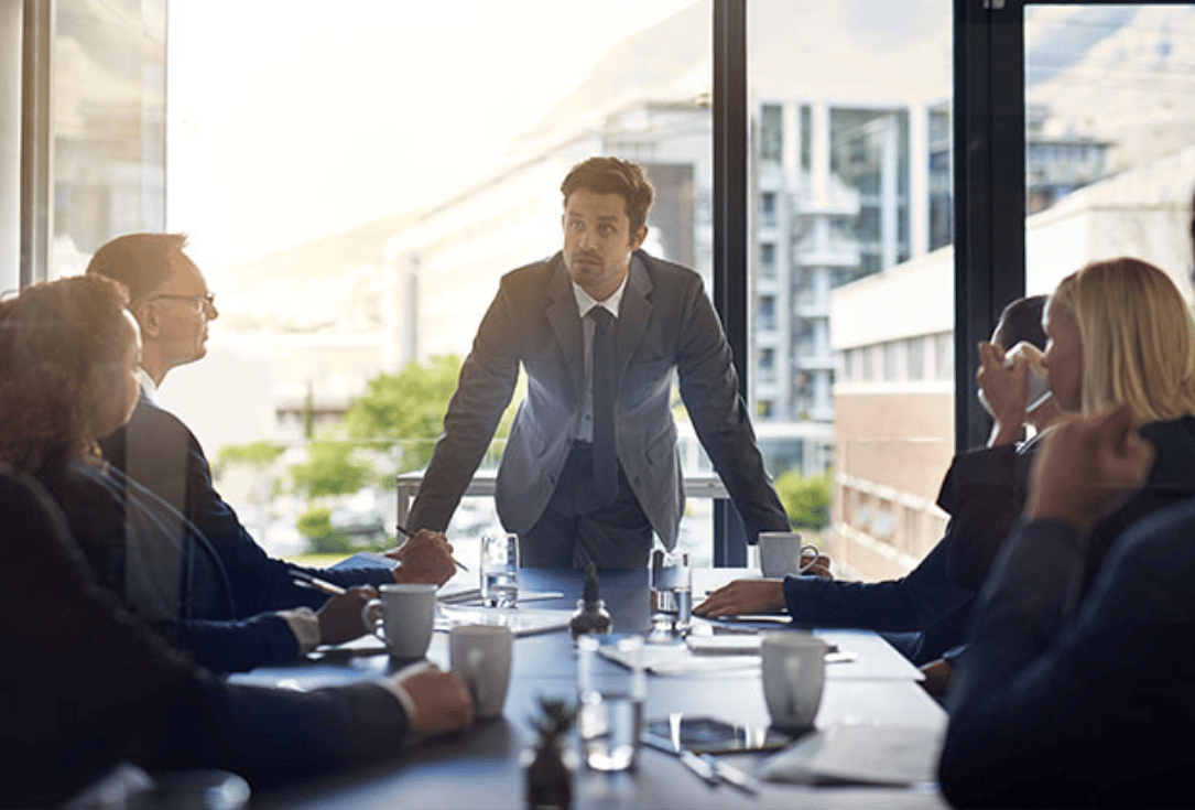 Man in a suit stands at a conference table addressing colleagues in a sunlit office setting.
