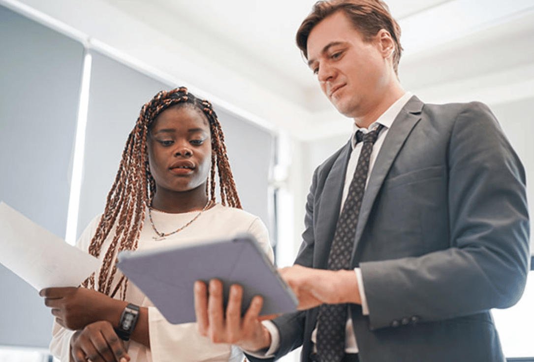 Two professionals review documents together in a bright office; a woman with braided hair holds papers while a man in a suit looks at a tablet.