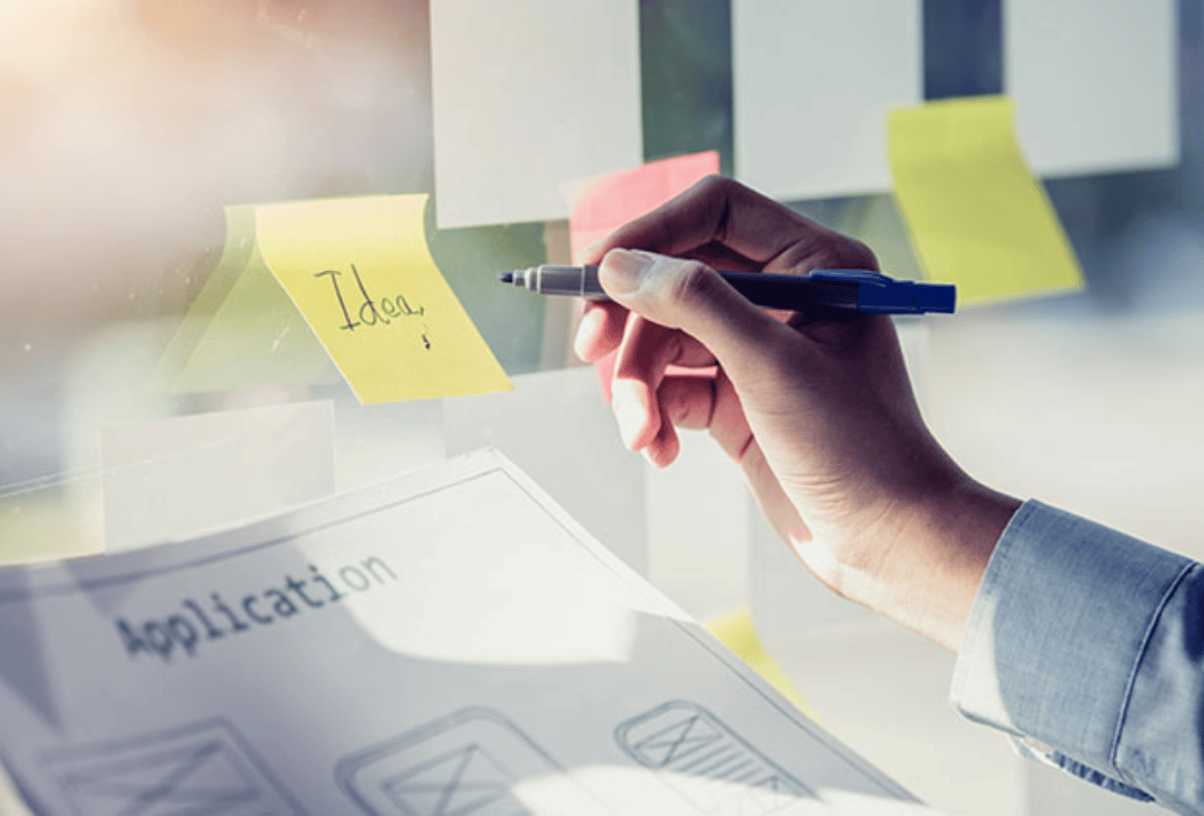 Close-up of a hand writing 'Idea' on a yellow sticky note attached to a glass board during brainstorming, with an 'Application' document below.