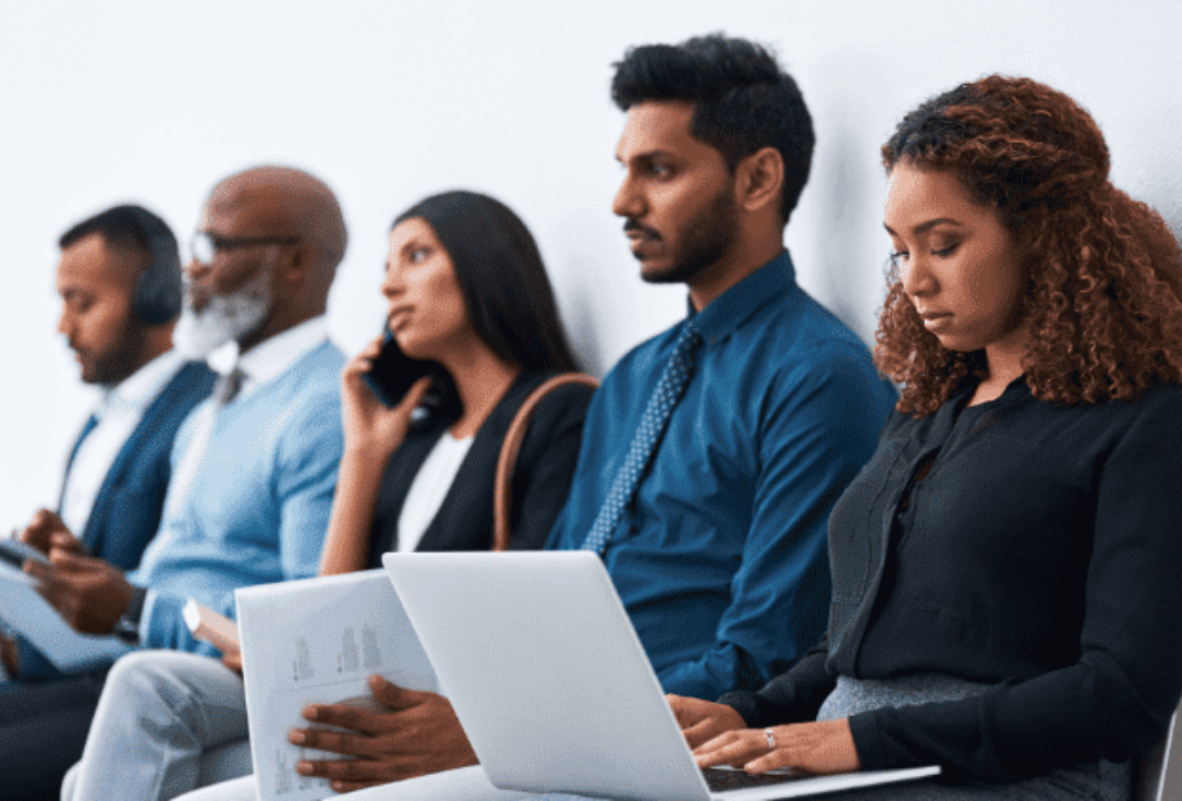 Diverse professionals seated in a row; foreground woman types on a laptop while others wait with papers or a phone.