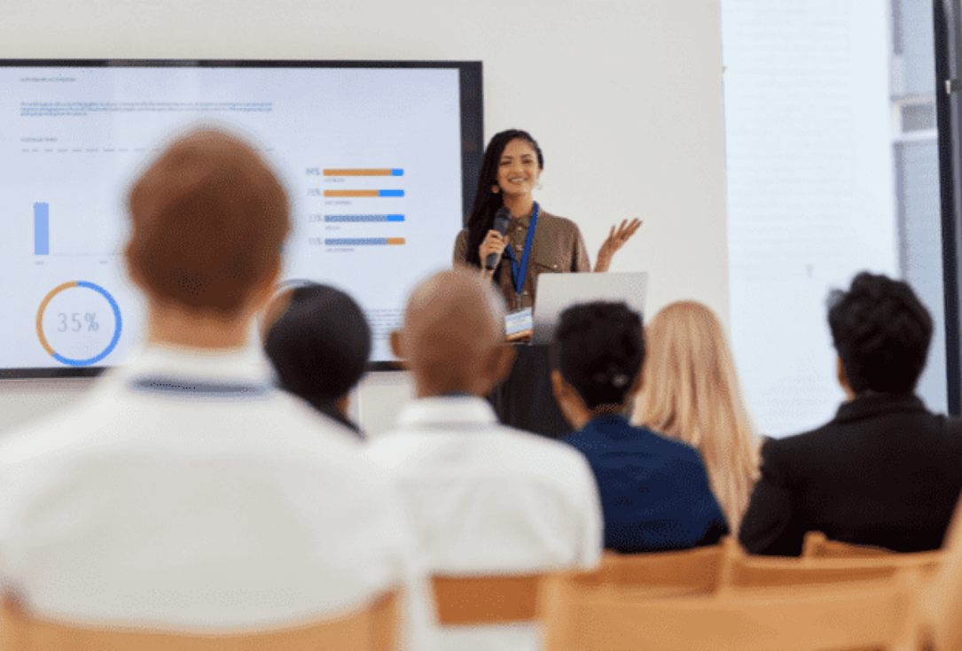 Female presenter with a microphone speaks to an audience beside a laptop; slides show charts on a large screen.