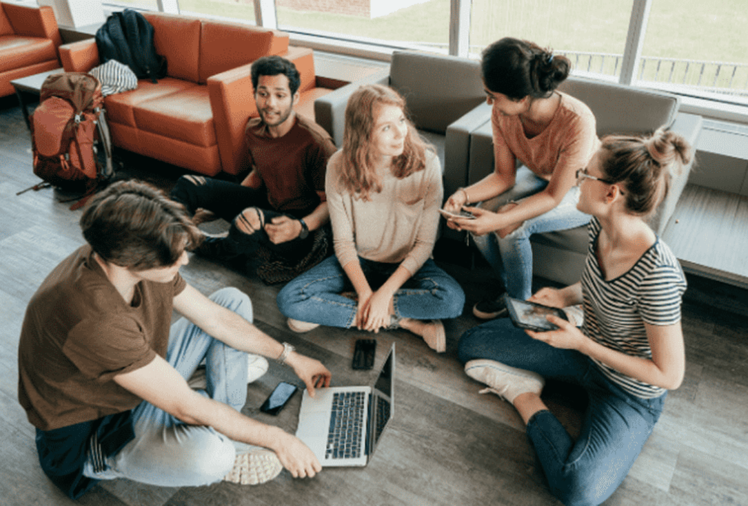 Five friends sitting cross-legged on the floor in a bright lounge, using laptops and phones to work together.
