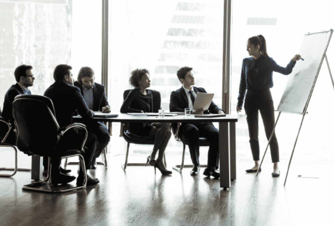 Business meeting in a modern office: a woman stands at a flip chart presenting to seated colleagues at a table of documents and laptops.