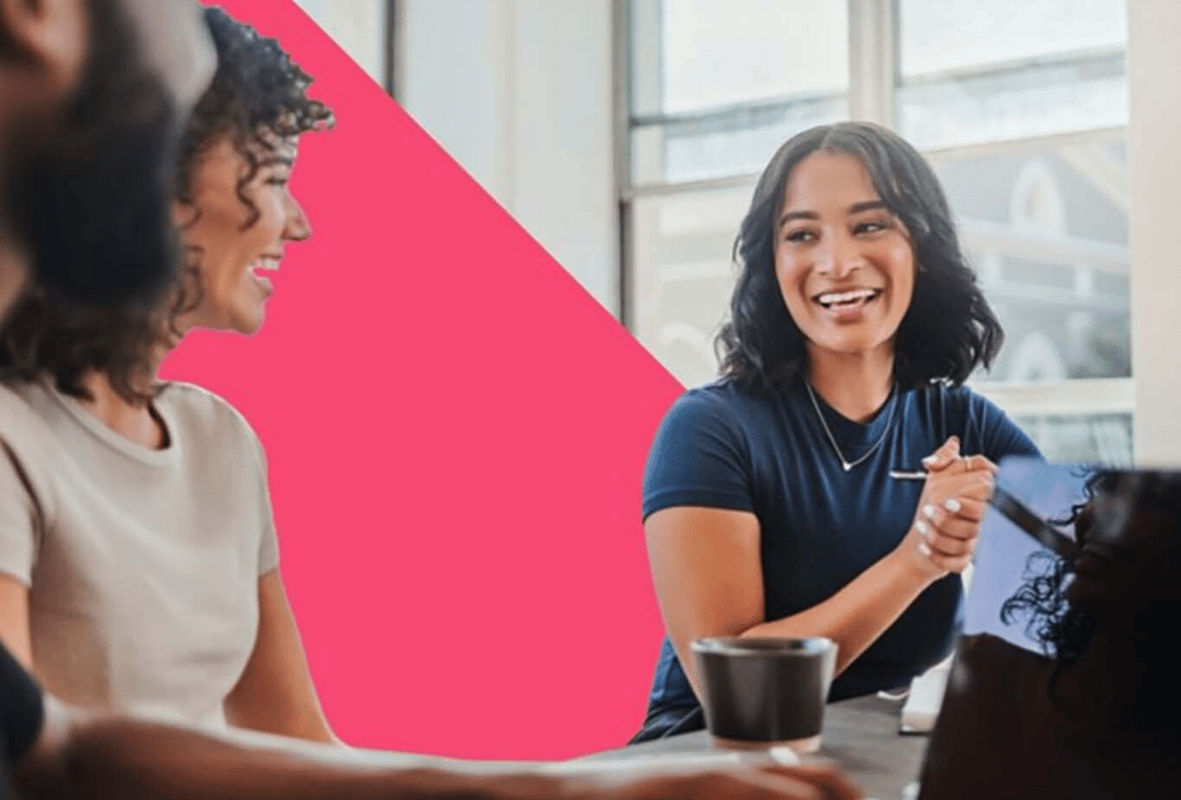 Two women in a bright office laugh and chat around a table during a meeting, with a bold pink diagonal backdrop in the background.