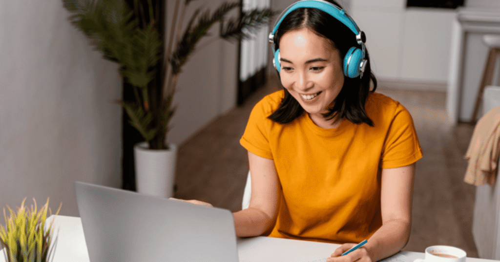 Woman wearing turquoise headphones smiling while using a laptop at a bright desk, taking notes with a pen.