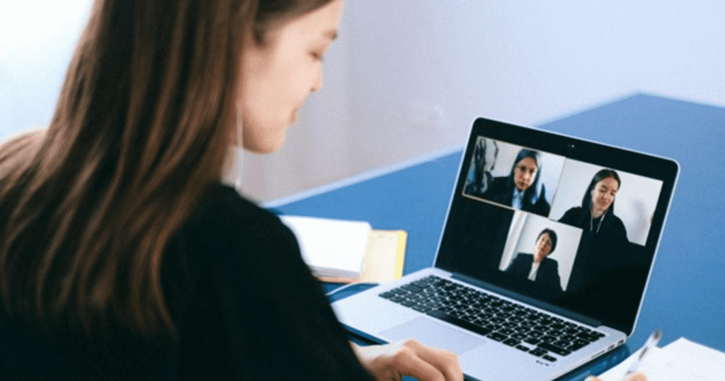 Person participating in a video conference on a laptop, four participants visible on screen, office/desk setting
