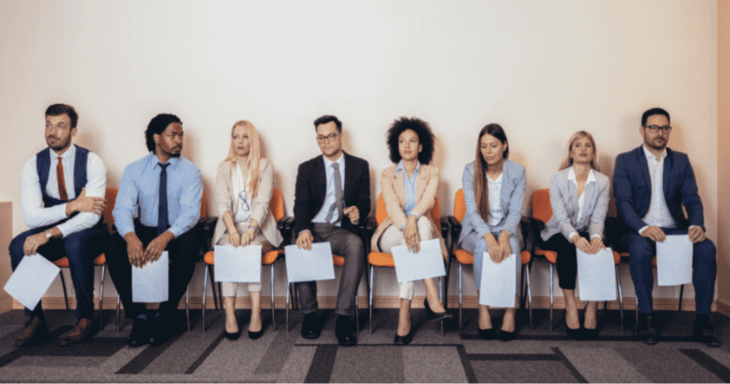 Group of seven professionals sitting in a row in a waiting area, each holding papers, dressed in business attire.