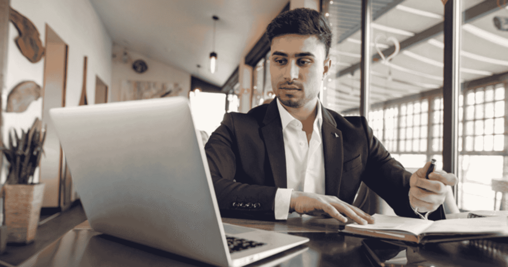 Man in a dark suit reviews documents at a desk with a silver laptop in a bright, modern office.