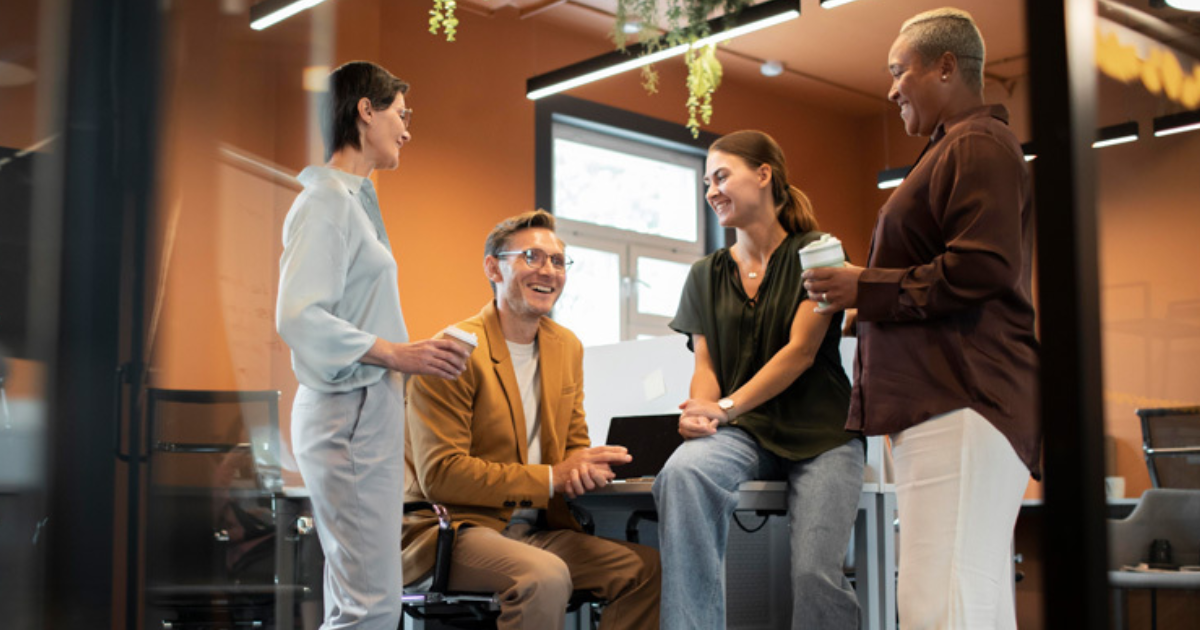 Four coworkers in a modern office chat around a laptop, holding coffee cups and smiling at each other for a collaborative discussion.