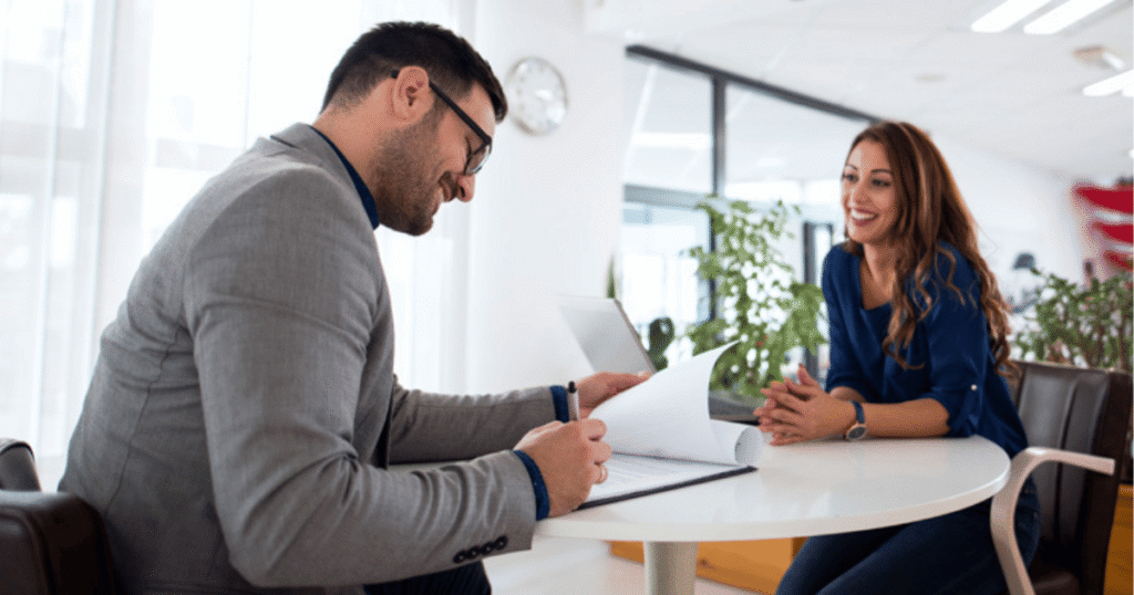 Man signs documents at a round white table while a smiling woman in blue watches in a bright office.