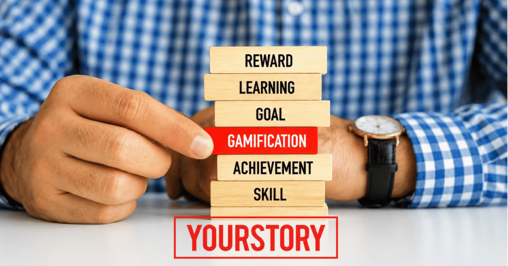 Close-up of a person pulling a red wooden block labeled "Gamification" from a stack of blocks labeled Reward, Learning, Goal, Achievement, and Skill, symbolizing gamification as a driver of learning, motivation, and achievement.