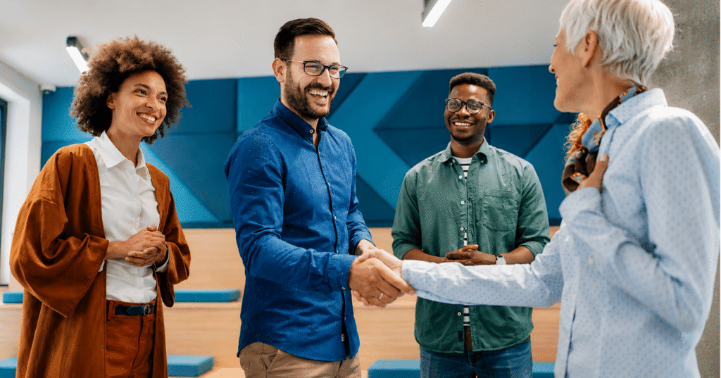A diverse group of professionals smiling and shaking hands in a modern office, symbolizing a successful meeting, collaboration, and a positive workplace culture.