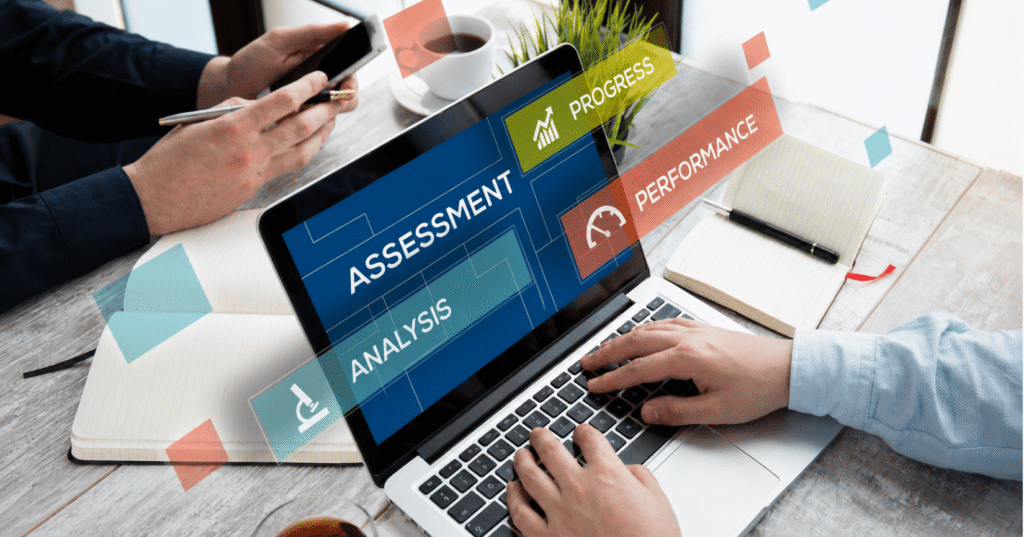 A professional office workspace with a laptop displaying digital assessment, analysis, progress, and performance dashboards, as two people collaborate using a smartphone, notebooks, and coffee at a modern desk.