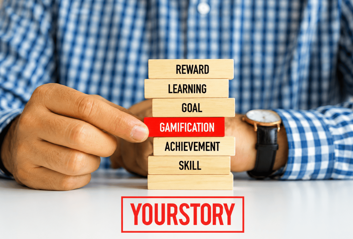 Close-up of a person pulling a red wooden block labeled "Gamification" from a stack of blocks labeled Reward, Learning, Goal, Achievement, and Skill, symbolizing gamification as a driver of learning, motivation, and achievement.