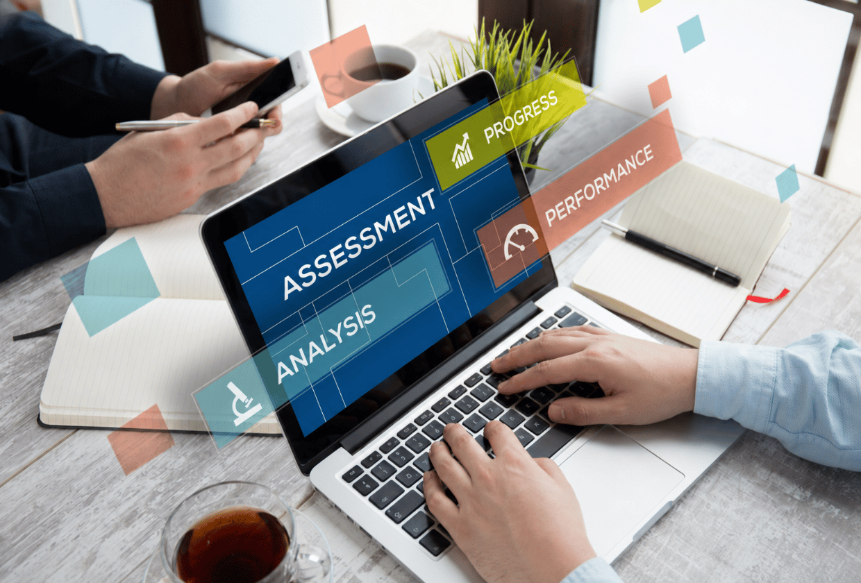 A professional office workspace with a laptop displaying digital assessment, analysis, progress, and performance dashboards, as two people collaborate using a smartphone, notebooks, and coffee at a modern desk.