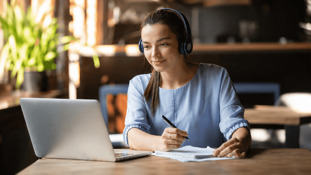 Young woman wearing headphones attends an online session on a laptop while taking notes at a desk in a modern workspace.