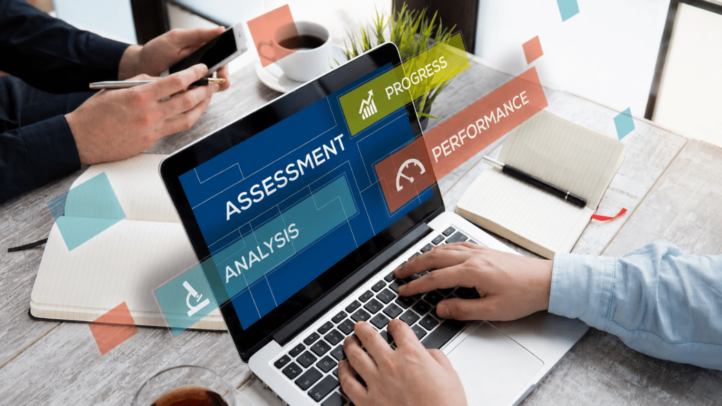 A professional office workspace with a laptop displaying digital assessment, analysis, progress, and performance dashboards, as two people collaborate using a smartphone, notebooks, and coffee at a modern desk.