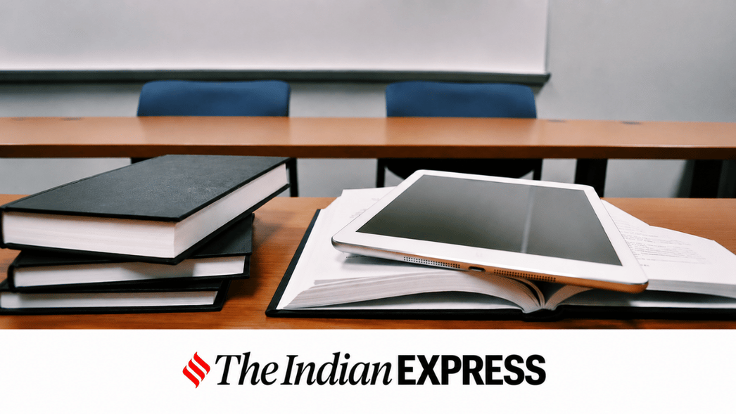Desk with stacked books, an open notebook, and a tablet in the foreground, with a blurred lecture room and The Indian Express logo at the bottom.
