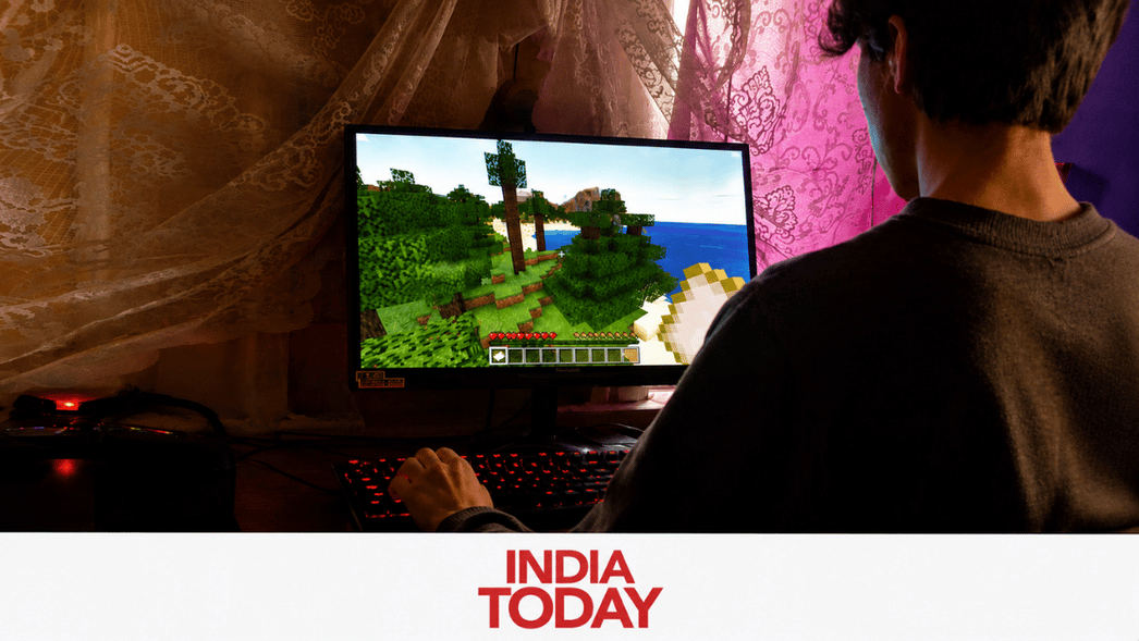 Person playing Minecraft on a desktop PC, seated at a desk with a lace curtain backdrop; monitor shows a grassy blocky landscape and a character’s hand visible on the right.