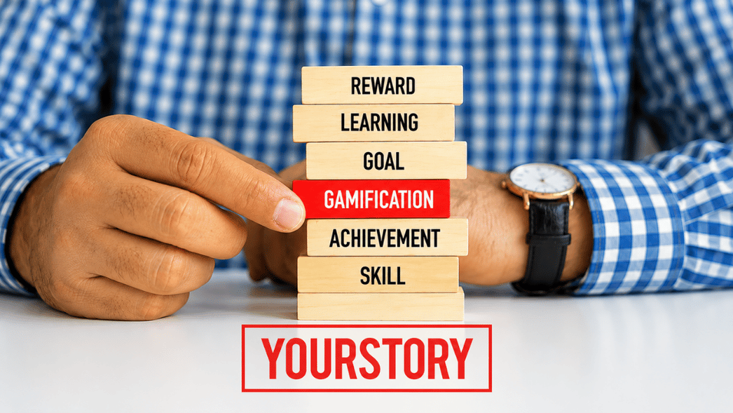 Close-up of a person pulling a red wooden block labeled "Gamification" from a stack of blocks labeled Reward, Learning, Goal, Achievement, and Skill, symbolizing gamification as a driver of learning, motivation, and achievement.