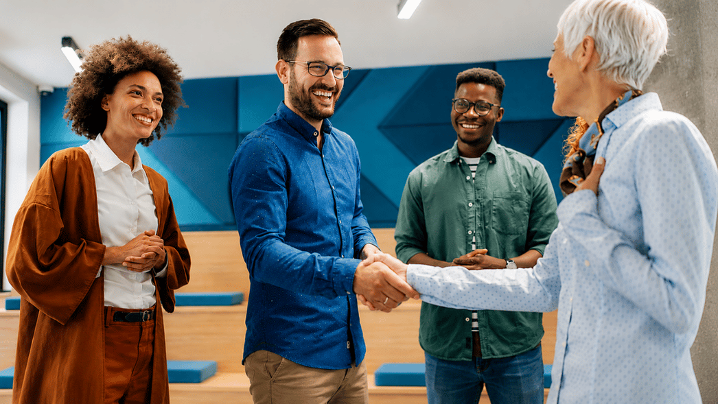 Four colleagues in a bright office smile as one man shakes hands with an older woman while two others look on.