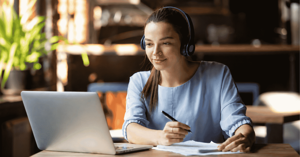Young woman wearing headphones attends an online session on a laptop while taking notes at a desk in a modern workspace.