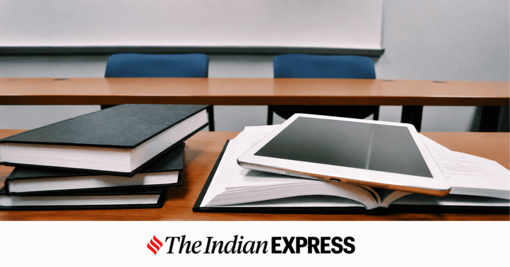 Desk with stacked books, an open notebook, and a tablet in the foreground, with a blurred lecture room and The Indian Express logo at the bottom.