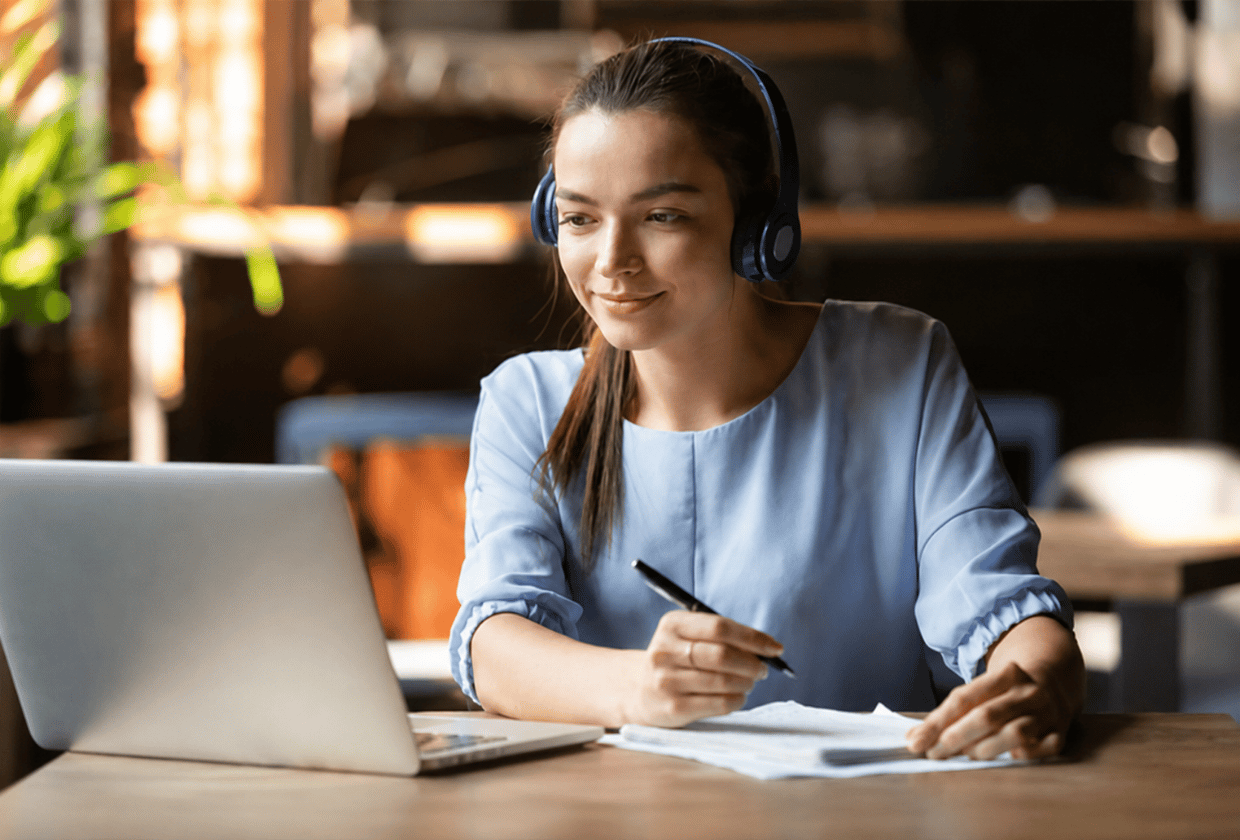 Young woman wearing headphones attends an online session on a laptop while taking notes at a desk in a modern workspace.