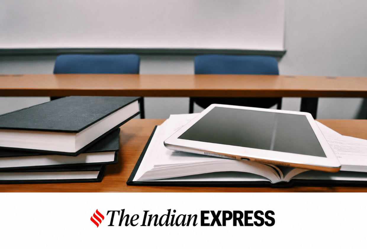 Desk with stacked books, an open notebook, and a tablet in the foreground, with a blurred lecture room and The Indian Express logo at the bottom.