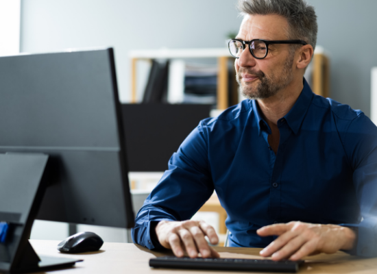Man in a blue shirt and glasses sits at a desk, working on a computer in a bright office environment.